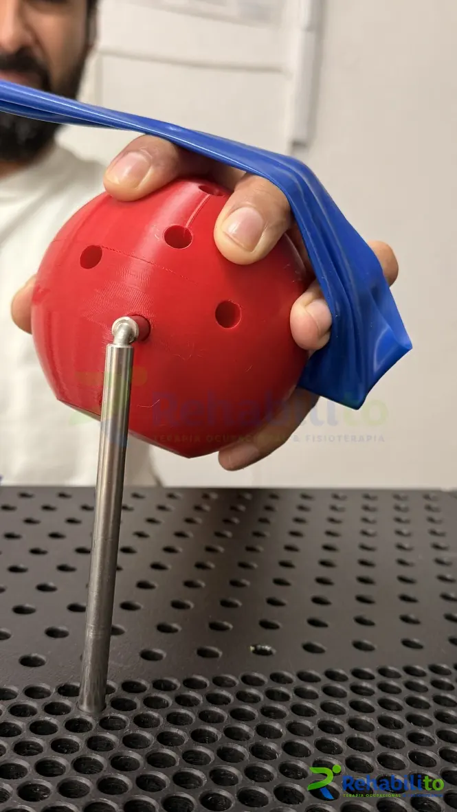Mano sujetando una esfera roja impresa en 3D con una banda elástica azul, ejercicio de prensión en terapia de mano